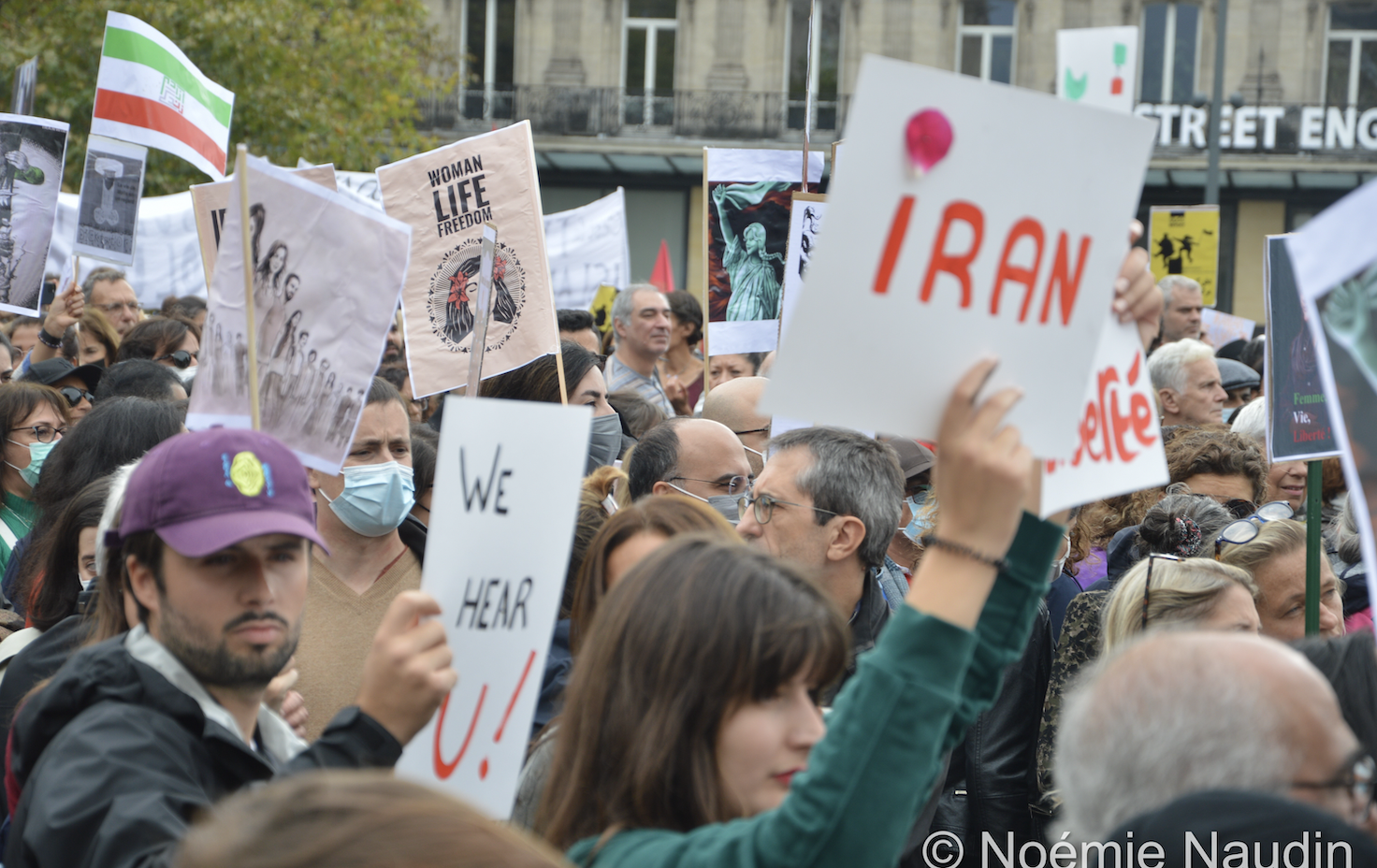 Une foule de personnes vue de côté, certaines tiennent des pancartes. Un homme et une femme au premier plan tiennent des pancartes disant "We Hear U!" (Nous vous entendons!) et "Iran" sur du papier blanc.